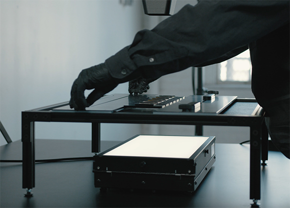 A person wearing black gloves and a dark long-sleeved shirt works with a black electronic device on a stand.