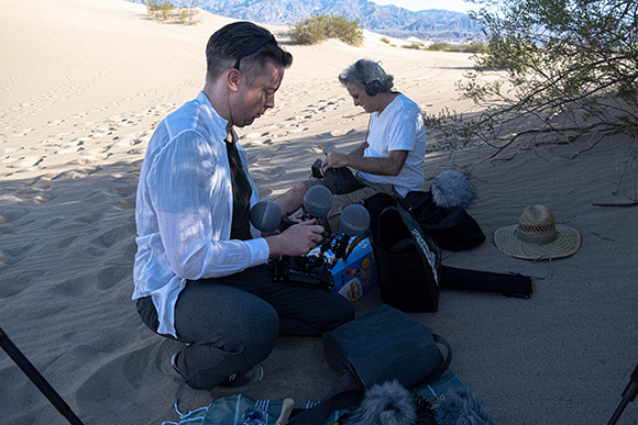 Two people crouch in sand dunes near desert shrubs, adjusting microphones and camera gear.