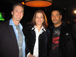A man in a blue shirt and blazer, a woman in a dark jacket, and a man in a black graphic t-shirt smiling.