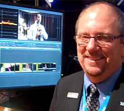 Smiling man with glasses and a suit standing in front of a monitor showing video editing software.