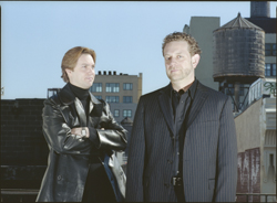 Two men in dark clothing pose on a rooftop against a city skyline and a wooden water tower.