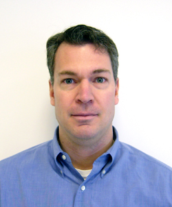 A man with short brown hair wearing a blue button-down shirt against a white background.