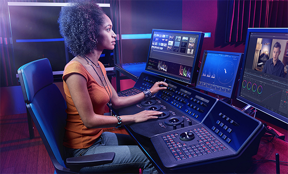 A woman sits at a large video editing desk with multiple monitors and control panels in a dark studio.