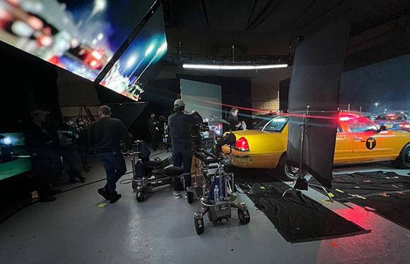 Film crew filming a yellow taxi cab on a studio set with large lighting screens and equipment.
