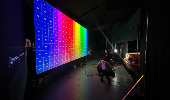 A large LED screen shows a grid of colorful squares ranging from blue to red in a dark studio setting.