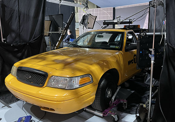 A yellow sedan taxi positioned in a film studio surrounded by lighting equipment and black curtains.