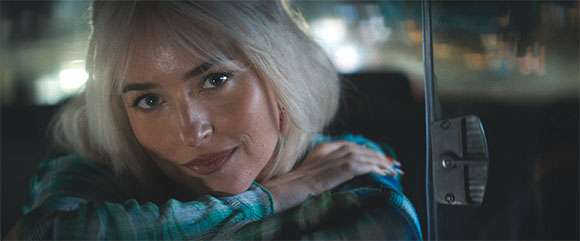 A blonde woman with a slight smile leans her chin on her hand inside a vehicle at night.