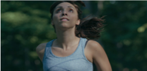 A woman in a grey tank top looks up toward the sky while running through a wooded area.