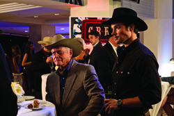 A man in a cowboy hat and suit sits near a man in a black cowboy hat and shirt.