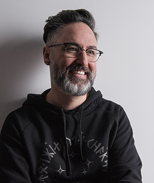 A man with short dark hair, glasses, and a grey-flecked beard smiling against a white wall.