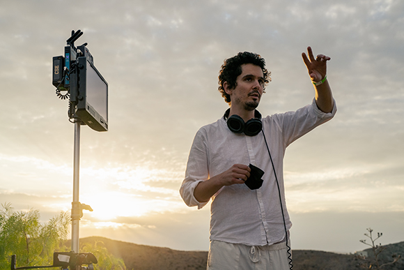 A man wearing headphones and a white shirt gestures toward the distance during a sunset film shoot.