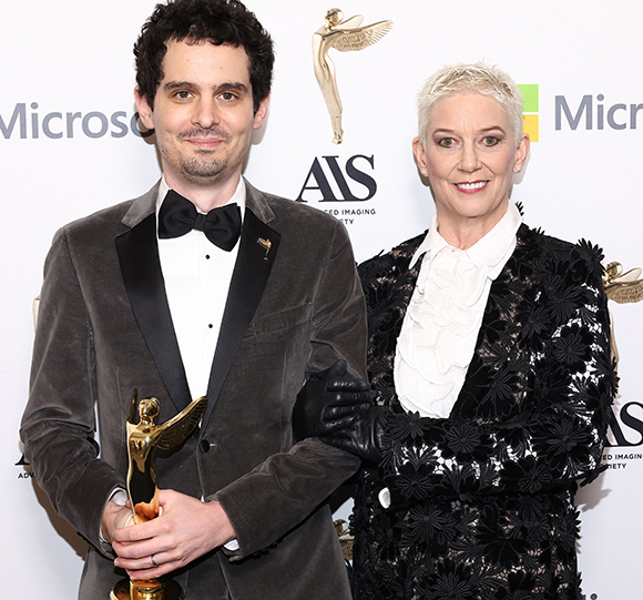 A man in a dark velvet tuxedo holding a gold award stands next to a woman with short blonde hair.