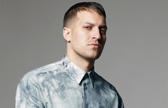 A man with short dark hair wearing a grey tie-dye patterned button-down shirt against a grey background.