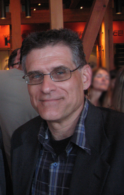 A man wearing glasses and a dark jacket over a plaid shirt smiles.
