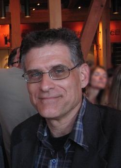 A man with short dark hair and glasses smiling, wearing a plaid shirt and dark blazer.