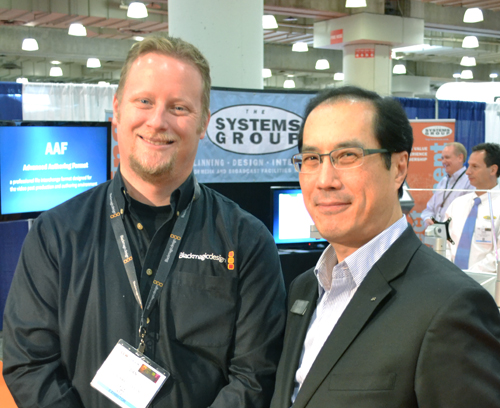 Two men smiling in front of a Systems Group booth at a convention.