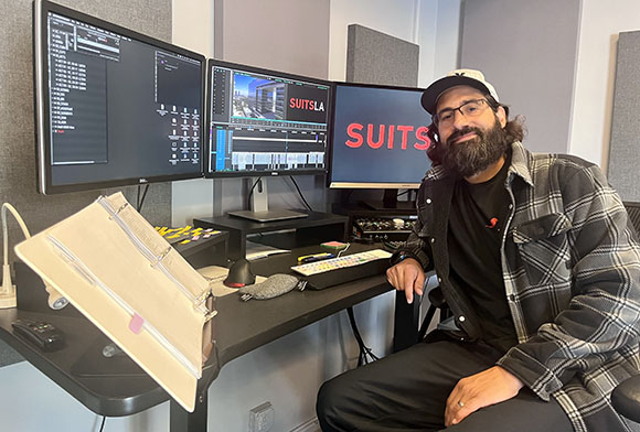 A bearded man in a plaid shirt sits at a desk with three large computer monitors displaying video editing software.