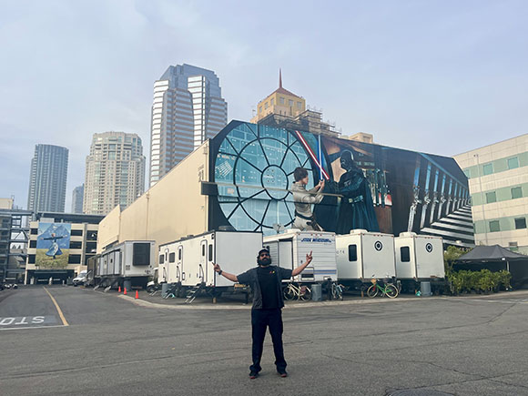 A man stands with arms outstretched in a parking lot before a large mural and city skyscrapers.