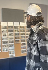 A man wearing a white baseball cap and black plaid shirt looks at a wall of small photographs.