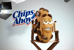 A personified chocolate chip cookie with eyes and teeth sitting in a director's chair next to a Chips Ahoy logo.