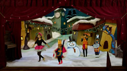 Performers in costumes dance on a snowy stage set with a snowman and winter village backdrop.