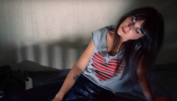 A woman with dark hair wearing a grey sleeveless shirt with a red heart design sits on a dark bed.