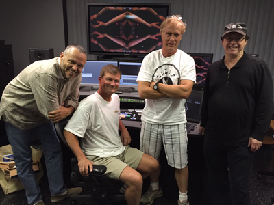Four men standing and sitting in a studio with computer monitors and audio equipment.