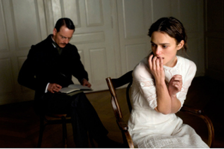A man in a dark suit reads a book in the background while a woman in a white dress looks anxious in the foreground.