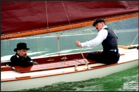 Two men in formal attire operating a small white sailboat with a large red sail on green water.