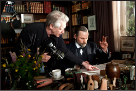 A man in a black shirt holds a camera while pointing at papers near another man in a suit.