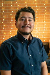 A man with dark hair and a goatee wearing a blue button-down shirt, smiling against a lit brick wall.