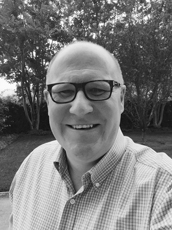 A smiling man with glasses and a checkered shirt outdoors with trees in the background.