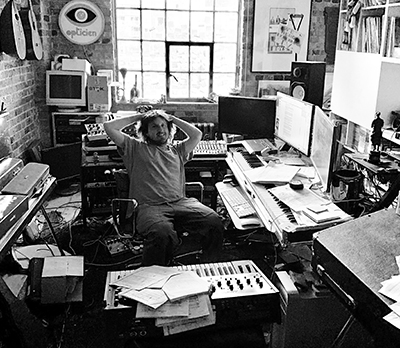 A man sits in a messy music studio filled with synthesizers, monitors, and audio gear.