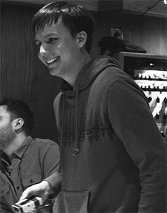 A smiling young man wearing a dark hoodie stands in a dimly lit room.