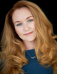 A woman with long, wavy reddish-blonde hair and blue eyes looks toward the camera against a black background.