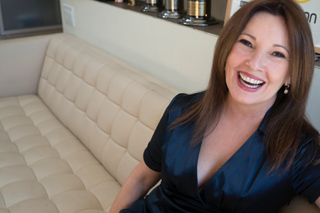 A smiling woman with long brown hair wearing a dark blue satin top sits on a beige tufted sofa.