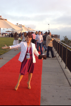 Woman in purple dress and white blazer on red carpet with arms outstretched, event in background.