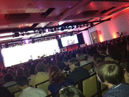 A large audience sits in a dimly lit conference hall, facing a brightly lit stage with two large screens. Red lighting illumi