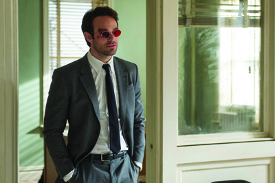 A man wearing a grey suit, white shirt, dark tie, and red-tinted sunglasses stands in a doorway.
