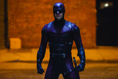 A person wearing a dark purple and black tactical superhero suit and mask standing in a dimly lit street.