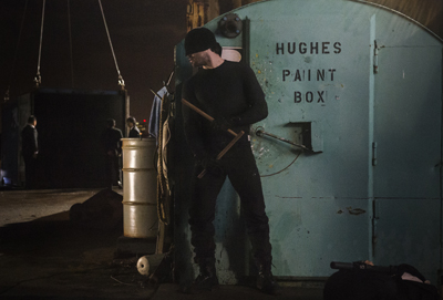 A man in black tactical gear stands before a large, weathered blue metal door in a dark industrial setting.
