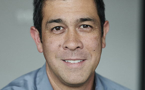 A close-up portrait of a man with short dark hair smiling against a neutral background.