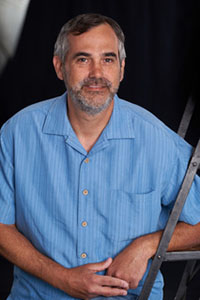 A man with graying hair and a beard wearing a light blue striped button-down shirt.