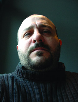 A man with a dark beard and a dark turtleneck sweater looks upward against a dark background.