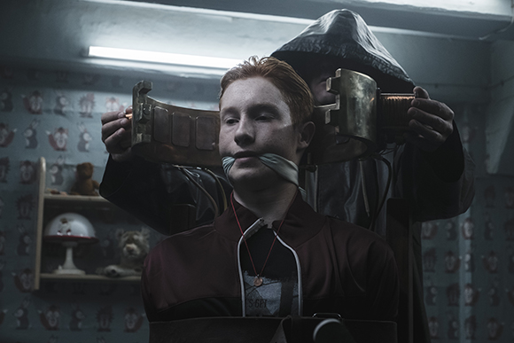 A person in a dark hood places a large metal collar around the head of a seated red-haired person.
