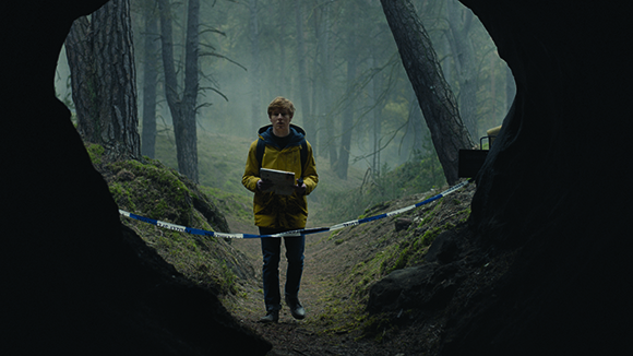 A person in a yellow jacket stands in a misty forest behind police tape, framed by a dark cave opening.