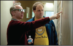 A man in a red sweater points at a wall while another man in a yellow shirt watches.