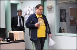 A man in a yellow shirt and blue jacket walks through an office past a man in a suit.