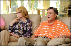 A woman with blonde hair and glasses sits next to a man in an orange striped polo shirt on a beige couch.