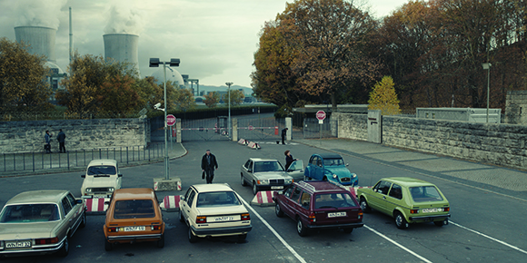 Vintage cars parked in a lot before a stone wall, with industrial cooling towers in the background.